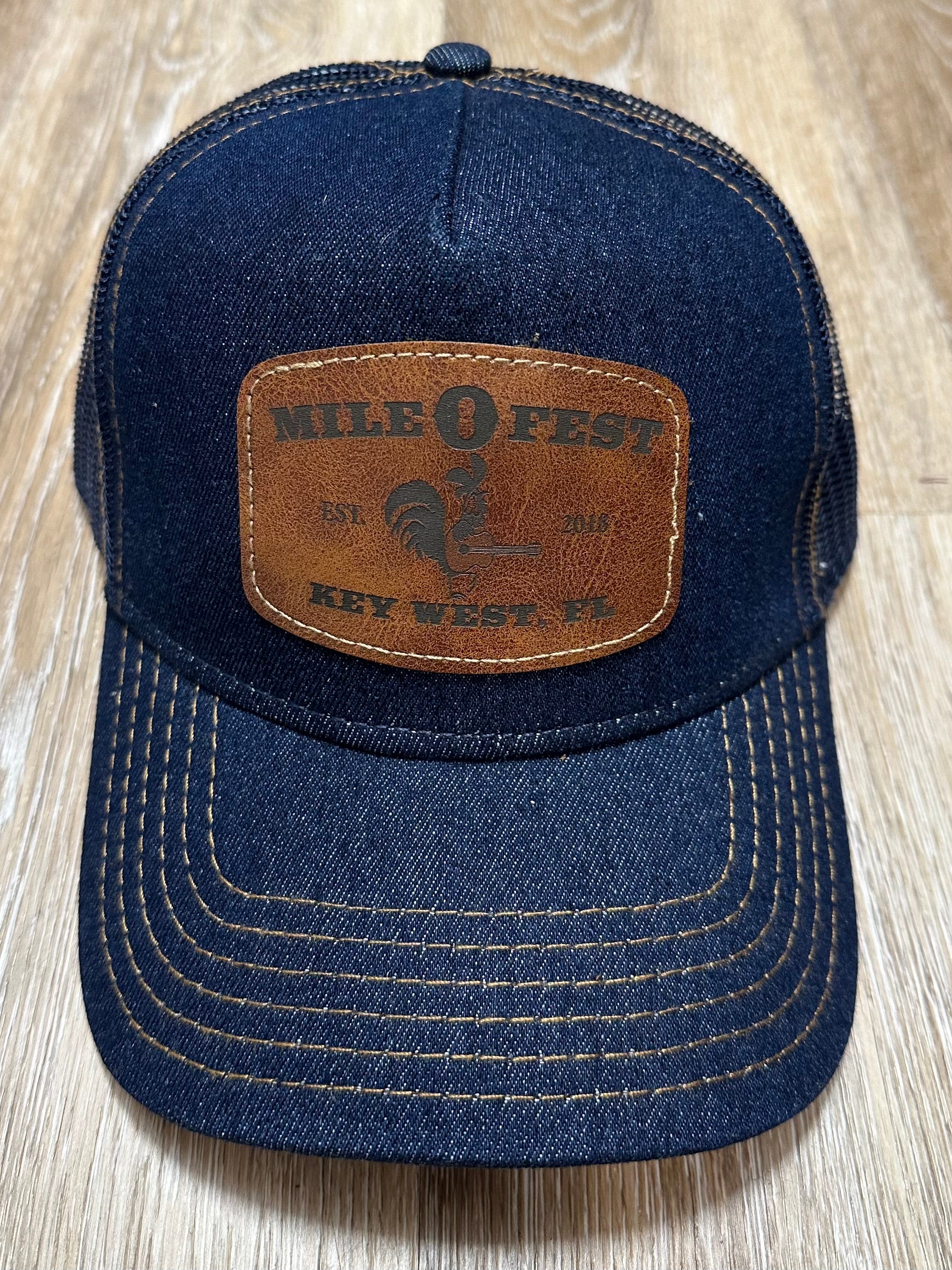A blue denim trucker hat with a leather patch on the front featuring text and a rooster illustration.